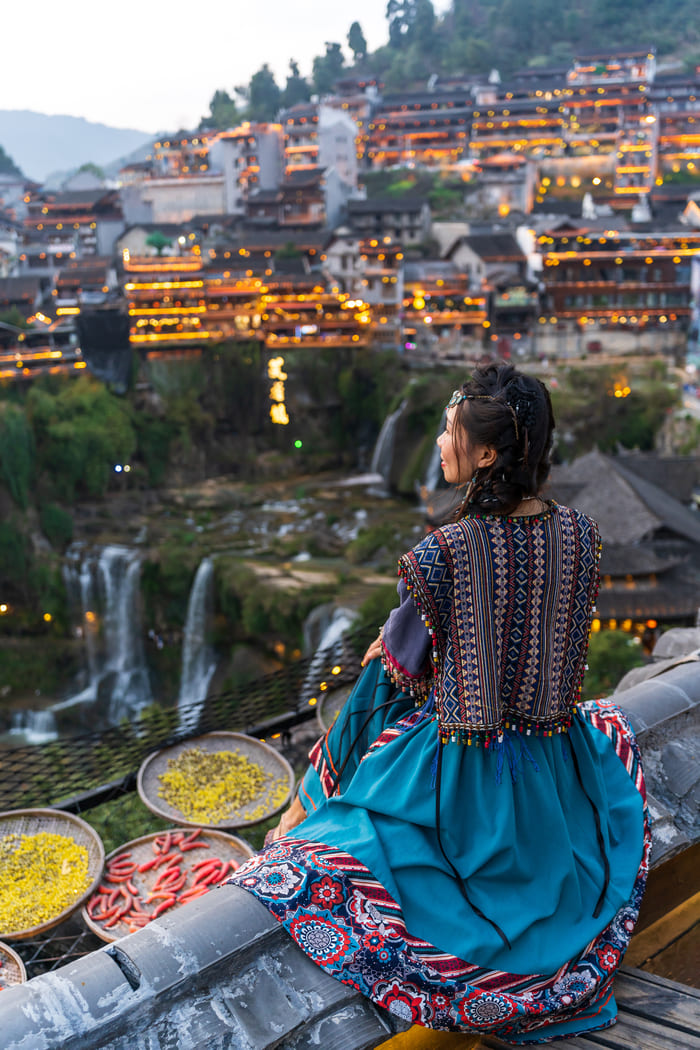 Primer plano de una mujer de espaldas con un colorido vestido étnico y un chaleco bordado, mirando hacia el casco antiguo de Lijiang al anochecer, que cuenta con edificios de madera tradicionales, luces cálidas y cascadas que caen en un valle.