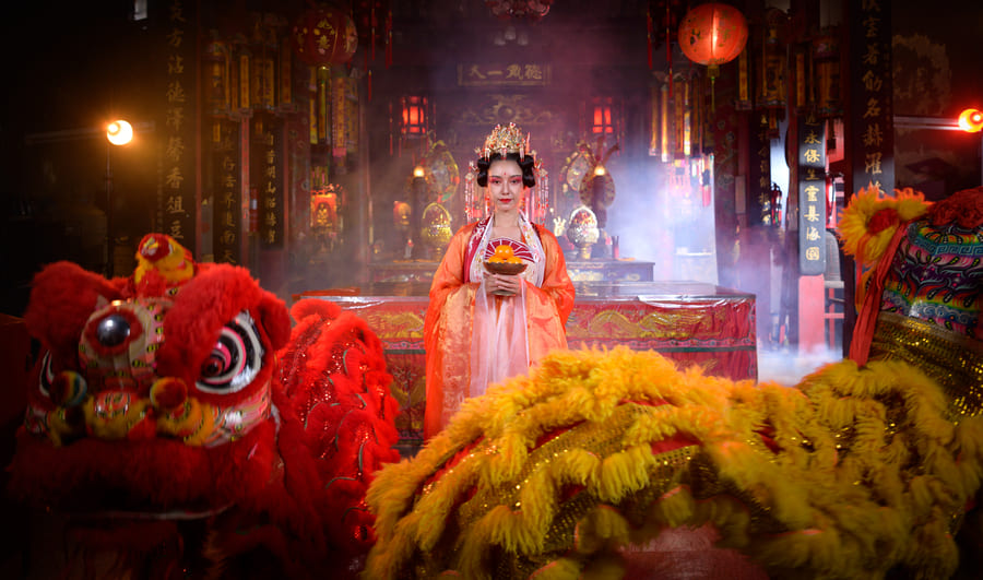 Una mujer con un elaborado traje tradicional chino de color naranja y un tocado dorado sostiene mandarinas dentro de un templo chino lleno de humo. En primer plano se aprecian dos coloridos disfraces de danza del león: uno rojo a la izquierda y otro amarillo a la derecha. El fondo muestra linternas rojas colgadas y elementos ceremoniales bajo una luz ambiental cálida.
