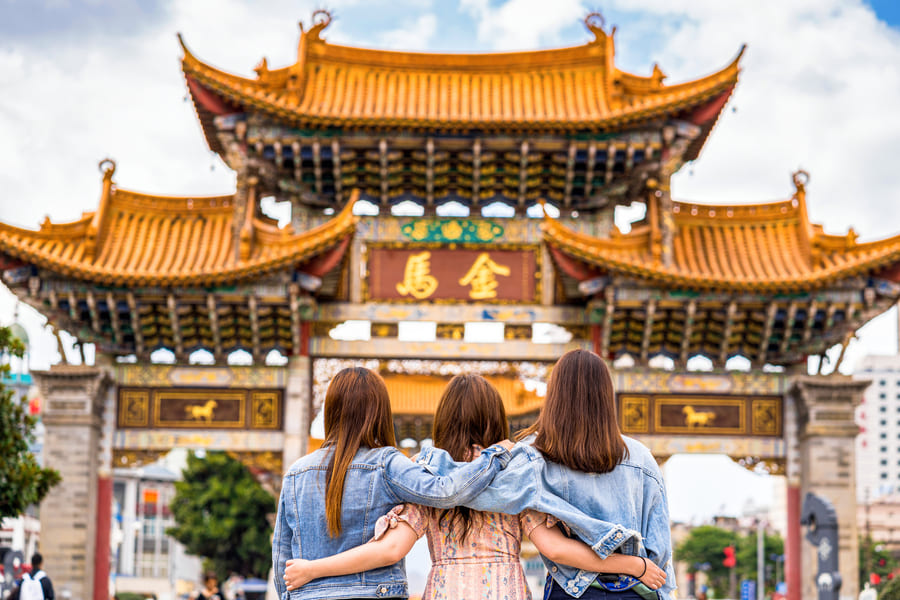 Vista desde atrás de tres mujeres jóvenes o amigas abrazadas por los hombros mientras contemplan una gran puerta monumental tradicional china (Paifang). Visten chaquetas de mezclilla y miran hacia la imponente estructura arquitectónica de color ocre y dorado, decorada con intrincados detalles y caracteres chinos en el centro, bajo un cielo parcialmente nublado.
