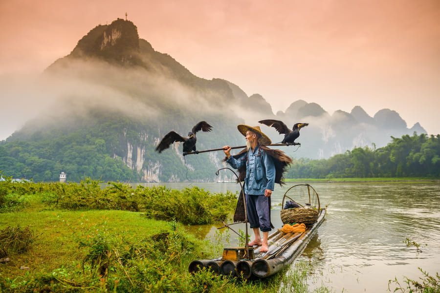 Un pescador tradicional chino con sombrero de paja y capa de lluvia está de pie sobre una balsa de bambú en el río Li en Yangshuo. Lleva una vara sobre los hombros con dos aves cormoranes con las alas abiertas. Al fondo, se aprecian las icónicas colinas cársticas rodeadas de niebla bajo un cielo con tonos rojizos al amanecer.