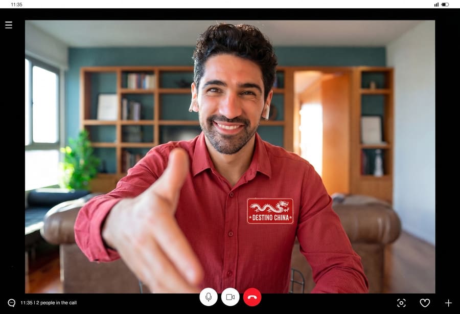 Un hombre sonriente en una videollamada extiende la mano en saludo, con un parche de "DESTINO CHINA" en su camisa roja.