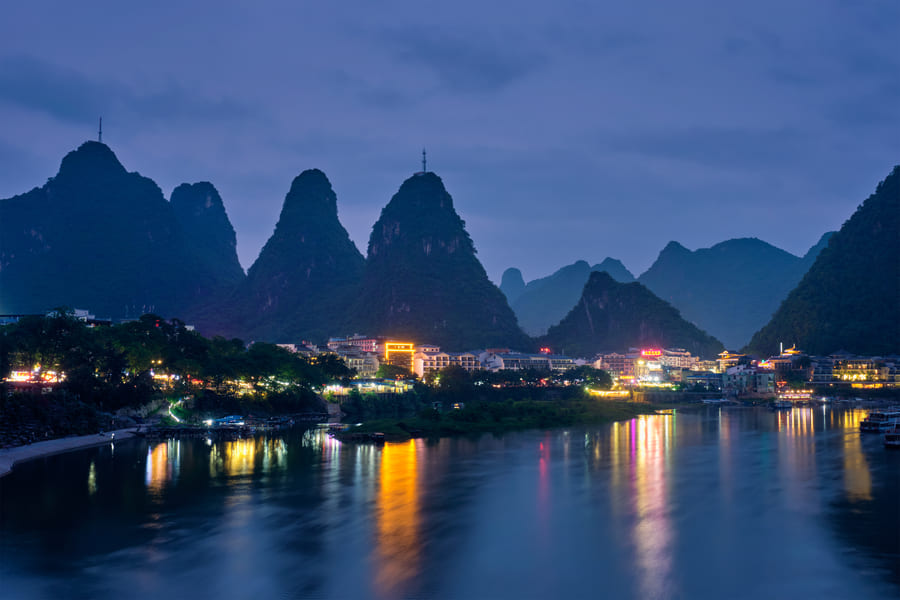 Paisaje nocturno de Yangshuo con luces de la ciudad reflejadas en el río Li y colinas kársticas iluminadas al fondo.