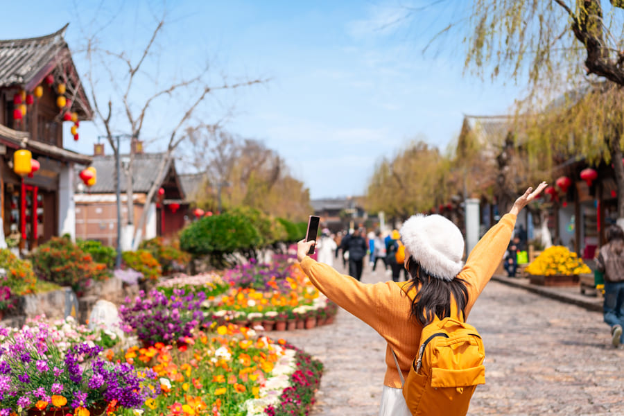 Turista con mochila amarilla fotografiando una calle peatonal decorada con flores y linternas tradicionales en un pueblo antiguo de Yunnan, China.